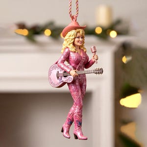 Op de afbeelding: Een hangend ornament met een blonde figuur in een roze cowboyhoed, een roze jumpsuit met vlinder- en bloemenpatronen en roze cowboylaarzen. De figuur houdt een zilveren microfoon en een roze akoestische gitaar met het woord DOLLY vast.