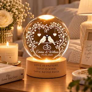 Könnte beinhalten: Ein kugelförmiges Glas-Nachtlicht auf einem zylindrischen Holzsockel. Das Glas zeigt ein geätztes Design mit zwei Vögeln auf einem Ast, Herzen, Eheringen und dem Text "Emma & William 25.06.2026". Der Holzsockel ist mit "LOVE IS PATIENT, LOVE IS KIND, LOVE NEVER FAILS." graviert.