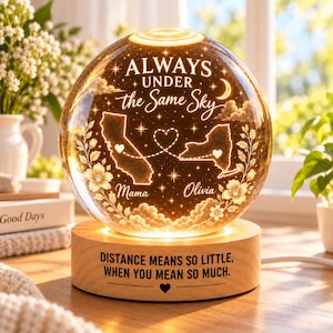 Op de afbeelding: Een bolvormig glazen nachtlampje op een cilindrische houten voet. Het glas bevat de tekst "ALWAYS UNDER the Same Sky", contouren van Californië en New York met hartjes, en de namen "Mama" en "Olivia". De voet bevat de tekst "DISTANCE MEANS SO LITTLE, WHEN YOU MEAN SO MUCH."