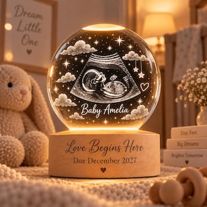 May include: A spherical glass night light featuring an engraved ultrasound image, clouds, stars, and the text Baby Amelia. The light sits on a cylindrical wooden base engraved with the text Love Begins Here and Due December 2027 with a heart icon.