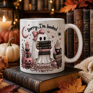 May include: A white ceramic mug featuring a ghost illustration with a pink bow, holding a stack of books labeled Bookish, Curious, Cozy, Imaginative, and Thoughtful. The text reads Sorry, I'm booked and Brianna, with pink pumpkins, candles, and stars.