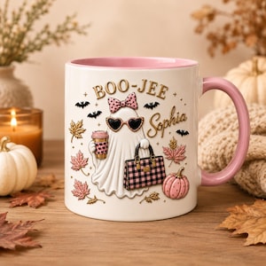 May include: A white ceramic mug with a pink interior and handle, featuring a ghost illustration wearing a pink polka-dot bow and heart-shaped sunglasses. The ghost holds a coffee cup and a pink plaid handbag. Text reads BOO-JEE and Sophia.