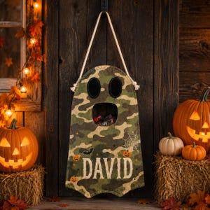 May include: A ghost-shaped wall hanging made of camouflage fabric with a white rope hanger. The ghost features black oval eyes, a circular mouth opening, and the name DAVID printed in white, surrounded by small orange pumpkins and a spiderweb.
