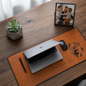 Op de afbeelding: Een bruine leren bureaumat met een zwarte rand, een laptop, muis en pen. De mat heeft een familiefoto en de tekst "Happy Father's Day". Een ingelijste foto en een vetplant in een geometrische pot staan ook op het houten bureau.