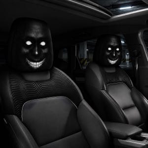 May include: Two black car headrest covers featuring a high-contrast graphic of a dark, shadowy face with glowing white eyes and a wide, toothy grin. The covers are fitted onto the front seats of a vehicle with black upholstery and textured panels.