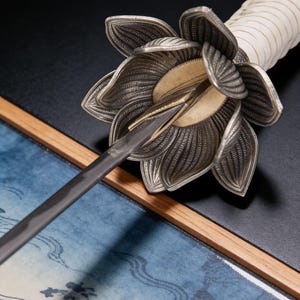 May include: Close-up of a decorative sword with a silver floral guard and a white wrapped handle. The blade is dark gray, and the guard has intricate petal details. The sword rests on a blue and white patterned surface.