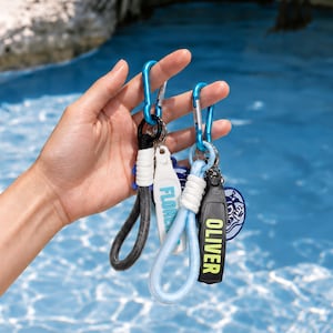 Può includere: Due portachiavi con moschettoni blu, cinturini da polso intrecciati in nero e azzurro, e targhette rettangolari bianche e nere. Una targhetta riporta il nome FLORENCE in blu, l'altra il nome OLIVER in giallo.