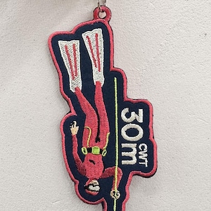 May include: A red and navy blue embroidered patch featuring a diver with the text "30m CWT". The diver is wearing a red suit, white fins, and a green mask. The patch is attached to a silver keyring.