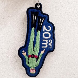 May include: A blue and green embroidered keychain featuring a diver in a green suit and black flippers, with the text "20m CWT". The diver is upside down, with arms outstretched. The keychain has a silver metal ring.