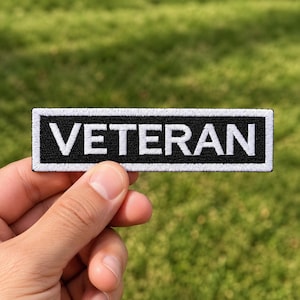 Könnte beinhalten: Ein rechteckiger, bestickter Aufnäher mit schwarzem Hintergrund und weißem Rand. Das Wort VETERAN ist in fetten, weißen Großbuchstaben in der Mitte des Aufnähers eingestickt. Das Design zeichnet sich durch eine klare, kontrastreiche Ästhetik mit sichtbarer Fadenstruktur auf der gesamten Oberfläche aus.