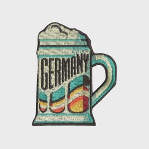 May include: Embroidered patch featuring a beer stein design with the word "GERMANY" in black. The stein is light blue with a white foam top and colorful accents of yellow, orange, and teal. The handle is teal.