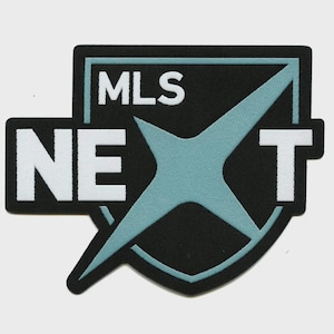 Op de afbeelding: Een zwart en blauwgroen MLS NEXT-patch. De patch heeft de tekst "MLS" boven een blauwgroen sterrenpatroon, met "NE" links en "T" rechts. De patch heeft een zwarte rand en staat tegen een witte achtergrond.