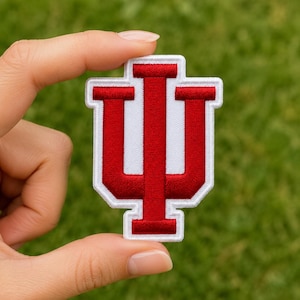 May include: A red and white embroidered patch featuring the Indiana University IU trident logo. The design consists of a bold red letter I integrated with a U shape, outlined in white thread with a clean, stitched border on a fabric base.