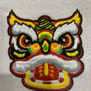 以下が含まれることがあります： 鮮やかな獅子舞の頭を刺繍したワッペン。赤、黄、白、黒の糸を使用し、目と口に焦点を当てています。服やアクセサリーに付けることを目的としたワッペンです。