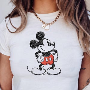 May include: White crop top featuring a black and red sketch of Mickey Mouse. The cartoon character is drawn with a pencil-like effect, with his signature red shorts and large white gloves. The top is a casual, everyday wear item.