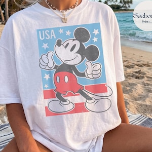 K&ouml;nnte beinhalten: Ein wei&szlig;es Kurzarm-T-Shirt mit einer Grafik von Mickey Mouse, die den Daumen hoch zeigt. Das Design enth&auml;lt den Text USA, wei&szlig;e Sterne auf hellblauem Grund und rote horizontale Streifen. Die Grafik hat ein abgenutztes Vintage-Finish.