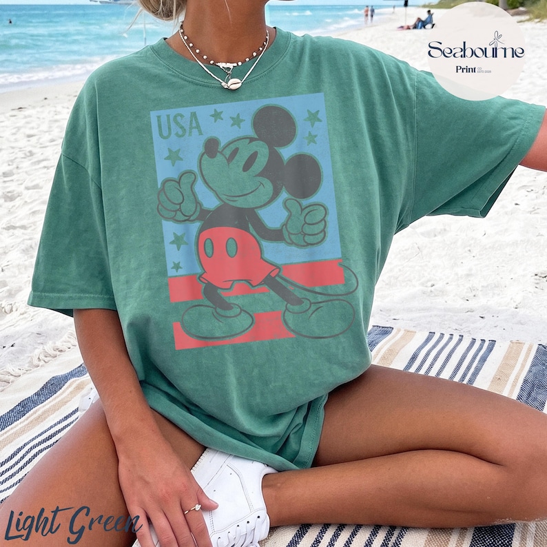 K&ouml;nnte beinhalten: Ein gr&uuml;nes kurz&auml;rmeliges T-Shirt mit einer Grafik von Mickey Mouse, die einen Daumen hoch zeigt. Das Design enth&auml;lt den Text USA, mehrere Sterne und rote horizontale Streifen. Das Shirt hat einen Rundhalsausschnitt und eine lockere Passform.