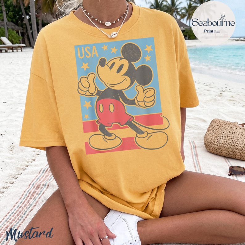 K&ouml;nnte beinhalten: Ein senfgelbes Kurzarm-T-Shirt mit einer Grafik von Mickey Mouse, die einen Daumen hoch zeigt. Das Design enth&auml;lt den Text USA, gelbe Sterne und rote Streifen auf einem hellblauen rechteckigen Hintergrund. Das T-Shirt hat einen Rundhalsausschnitt.