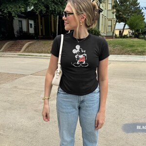 May include: Black t-shirt featuring a Mickey Mouse graphic in white and red. The shirt is paired with light blue jeans. The image is taken outdoors on a sunny day.