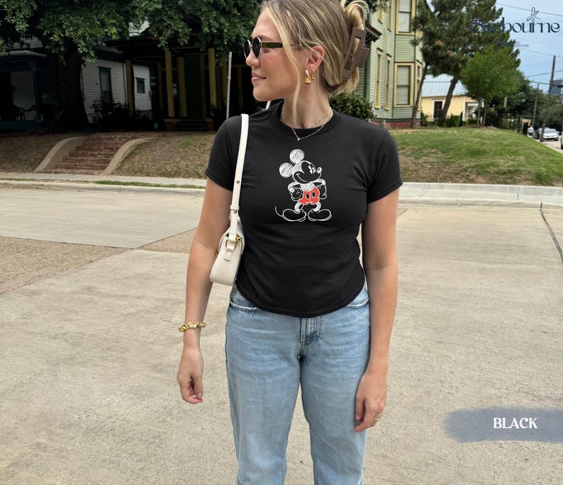 May include: Black t-shirt featuring a Mickey Mouse graphic in white and red. The shirt is paired with light blue jeans. The image is taken outdoors on a sunny day.