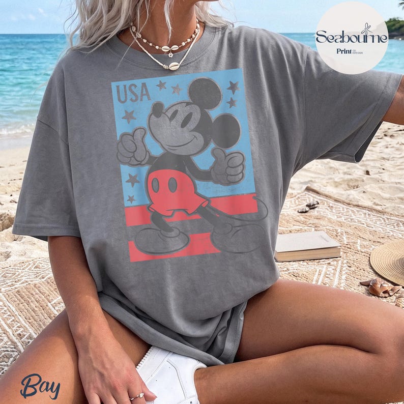 K&ouml;nnte beinhalten: Ein anthrazitfarbenes T-Shirt mit einer Grafik von Mickey Mouse, der beide Daumen nach oben zeigt. Das Design umfasst einen hellblauen Hintergrund, rote horizontale Streifen und kleine Sterne mit dem Text USA in der oberen linken Ecke.