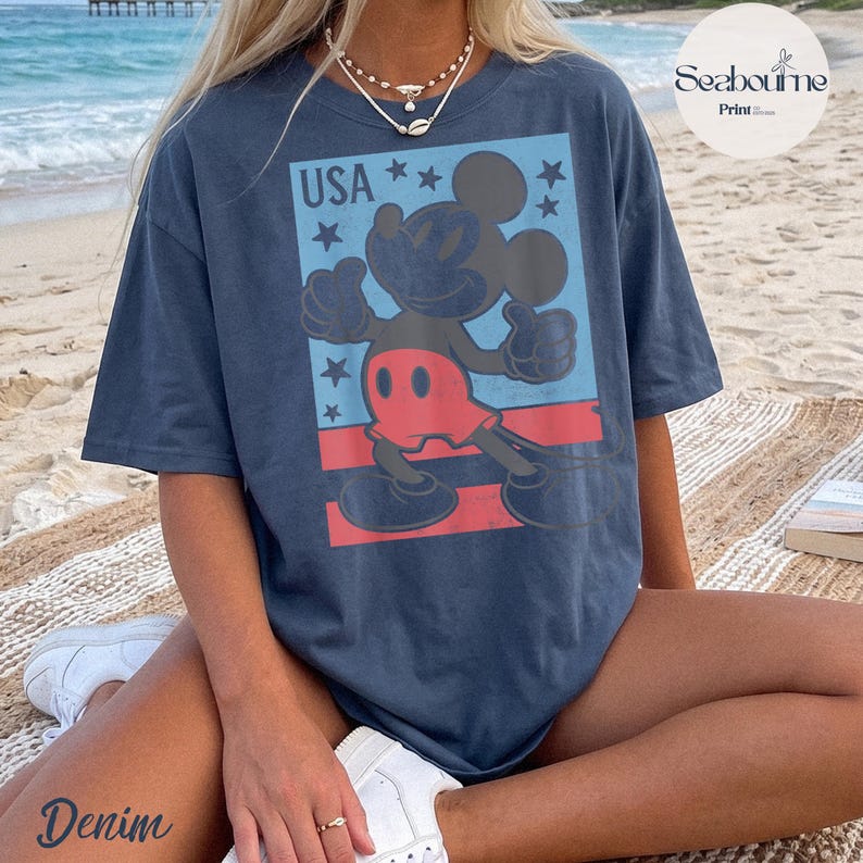 K&ouml;nnte beinhalten: Ein dunkelblaues Kurzarm-T-Shirt mit einem Grafikdruck von Mickey Mouse in roten Shorts, platziert vor einem hellblauen rechteckigen Hintergrund mit dem Text USA und mehreren dunkelblauen Sternen sowie roten Querstreifen am unteren Rand.