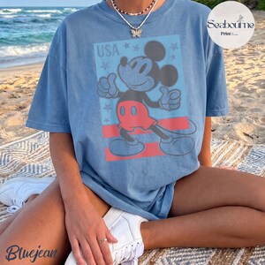 K&ouml;nnte beinhalten: Ein blaues Kurzarm-T-Shirt mit einem Grafikdruck von Mickey Mouse in roten Shorts und dem Text USA dar&uuml;ber. Das Design enth&auml;lt einen hellblauen rechteckigen Hintergrund mit roten horizontalen Streifen und kleinen Sternakzenten.