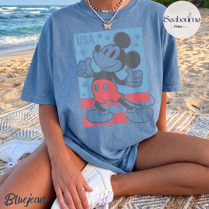 K&ouml;nnte beinhalten: Ein blaues Kurzarm-T-Shirt mit einem Grafikdruck von Mickey Mouse in roten Shorts und dem Text USA dar&uuml;ber. Das Design enth&auml;lt einen hellblauen rechteckigen Hintergrund mit roten horizontalen Streifen und kleinen Sternakzenten.