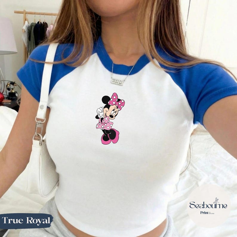 May include: White and blue raglan style t-shirt featuring a Minnie Mouse graphic. The cartoon character is wearing a pink polka dot dress and bow. The text "True Royal" is at the bottom left of the image.