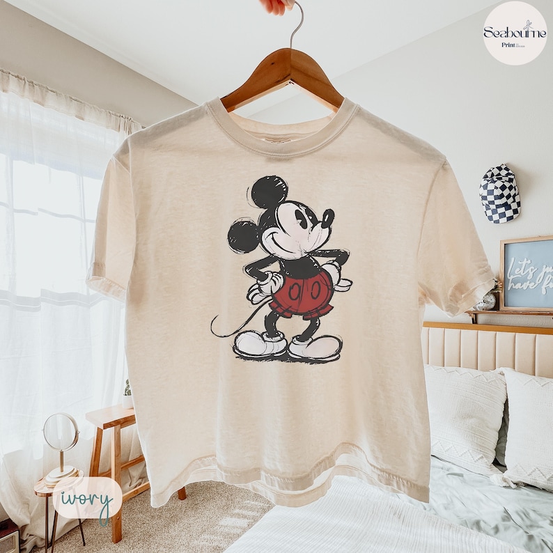 May include: Ivory-colored t-shirt featuring a distressed graphic of Mickey Mouse. The cartoon character is depicted in black, white, and red, with his hands on his hips. The shirt is hanging on a wooden hanger.