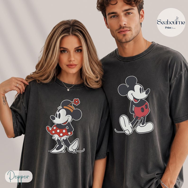 May include: Two charcoal gray t-shirts featuring Mickey and Minnie Mouse. Minnie Mouse is wearing a red polka dot dress and a flower hat. Mickey Mouse is wearing red shorts. The shirts are part of a matching set.