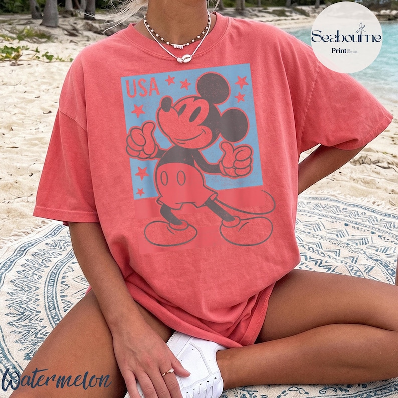 K&ouml;nnte beinhalten: Ein korallenfarbenes Kurzarm-T-Shirt mit einer Grafik von Mickey Mouse, die den Daumen hochh&auml;lt. Das Design enth&auml;lt einen hellblauen rechteckigen Hintergrund, rote Sterne und den Text USA. Das T-Shirt hat einen Rundhalsausschnitt und eine lockere Passform.