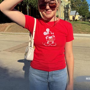 May include: A red t-shirt featuring a white graphic of Mickey Mouse. The shirt is short-sleeved and has a classic crew neck. The word "RED" is printed in white at the bottom right corner of the image. The shirt is paired with blue jeans.