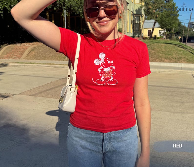 May include: A red t-shirt featuring a white graphic of Mickey Mouse. The shirt is short-sleeved and has a classic crew neck. The word "RED" is printed in white at the bottom right corner of the image. The shirt is paired with blue jeans.