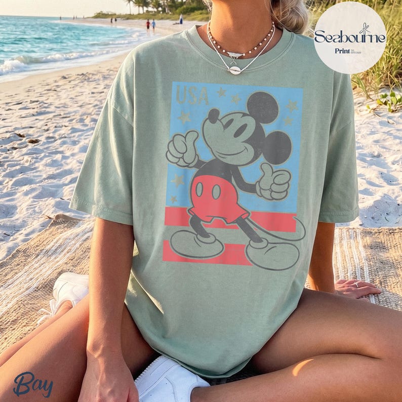 K&ouml;nnte beinhalten: Ein salbeigr&uuml;nes Kurzarm-T-Shirt mit einem Grafikdruck von Mickey Mouse, der einen Daumen hoch zeigt. Das Design enth&auml;lt den Text USA, Sterne und rote horizontale Streifen auf einem hellblauen rechteckigen Hintergrund. Rundhalsausschnitt.