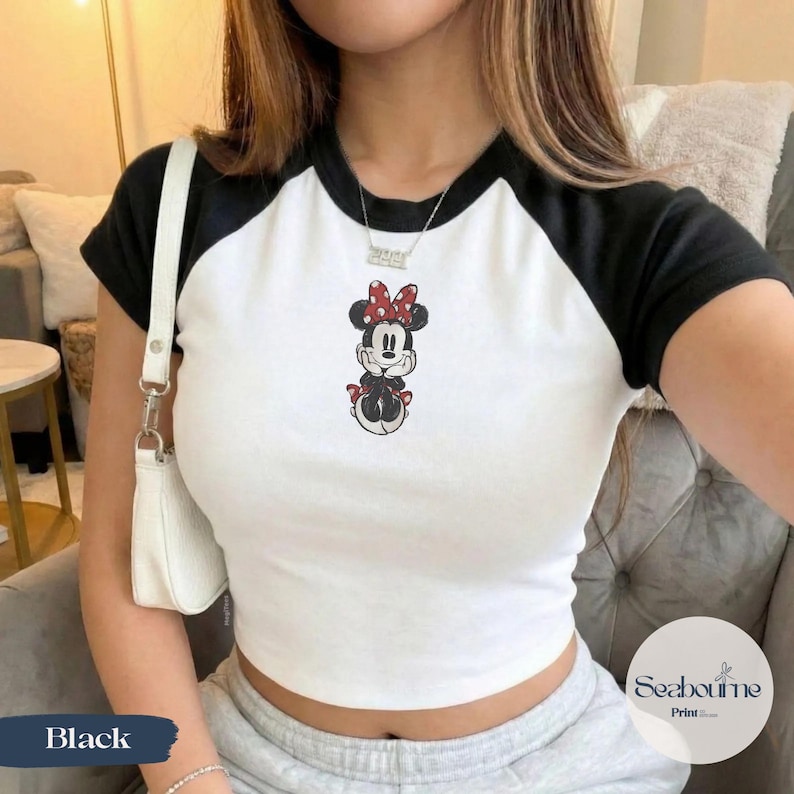 May include: White and black raglan style crop top featuring a Minnie Mouse graphic. The top has black sleeves and neckline trim. The Minnie Mouse graphic is in red, black, and white. The word "Black" is in the bottom left corner.