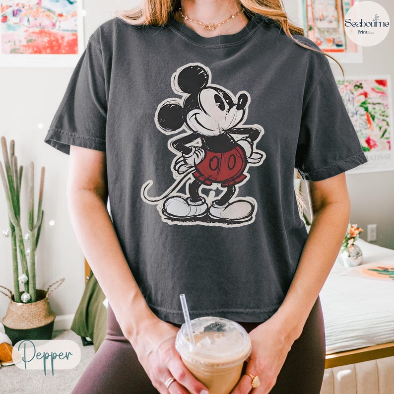 May include: Dark grey t-shirt featuring a distressed graphic of Mickey Mouse. The cartoon character is depicted in black, white, and red, with a classic pose. The shirt has short sleeves and a relaxed fit, suitable for casual wear.