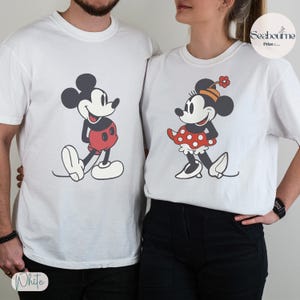 May include: White t-shirts featuring Mickey and Minnie Mouse. Mickey is in red shorts, and Minnie is in a red polka dot skirt. The shirts are printed with a vintage cartoon style.