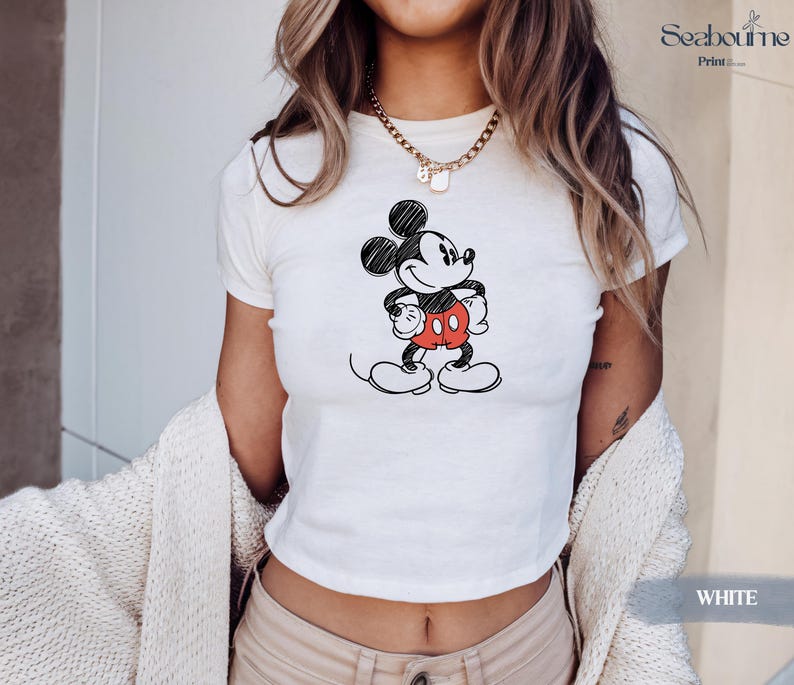 May include: White crop top featuring a black and red sketch of Mickey Mouse. The cartoon character is drawn with a pencil-like effect, with his signature red shorts and large white gloves. The top is a casual, everyday wear item.