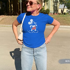 May include: Royal blue t-shirt featuring a Mickey Mouse graphic in white, red, and orange. The shirt is paired with light blue denim jeans. A white purse is worn over the shoulder.