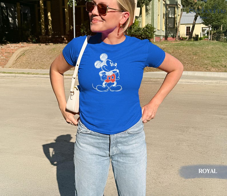 May include: Royal blue t-shirt featuring a Mickey Mouse graphic in white, red, and orange. The shirt is paired with light blue denim jeans. A white purse is worn over the shoulder.
