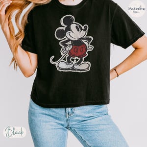 May include: Black t-shirt featuring a distressed graphic of Mickey Mouse in his classic pose. The cartoon character is depicted in black, white, and red. The shirt has short sleeves and a crew neck.