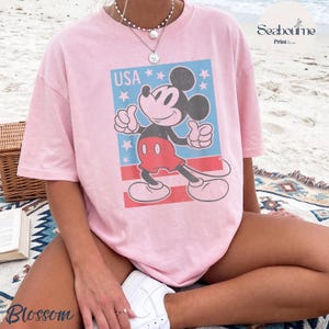 K&ouml;nnte beinhalten: Ein hellrosa T-Shirt mit kurzen &Auml;rmeln und einer Grafik von Mickey Mouse, der einen Daumen hoch zeigt. Das Design enth&auml;lt einen blauen rechteckigen Hintergrund mit wei&szlig;en Sternen, den Text USA und rote horizontale Streifen am unteren Rand.