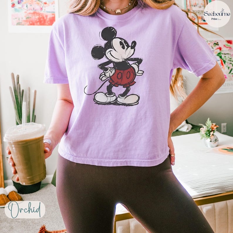 May include: A light purple t-shirt featuring a distressed graphic of Mickey Mouse in his classic pose. The cartoon character is wearing red shorts and white gloves and shoes. The word "Orchid" is printed on the bottom left of the image.