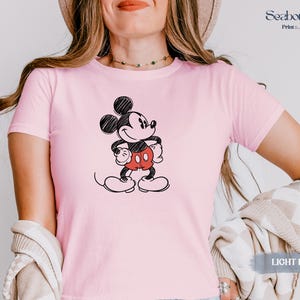 May include: Light pink t-shirt featuring a black and white sketch of Mickey Mouse with red shorts. The shirt has short sleeves and a crew neck. The text "LIGHT PINK" is visible in the bottom right corner.