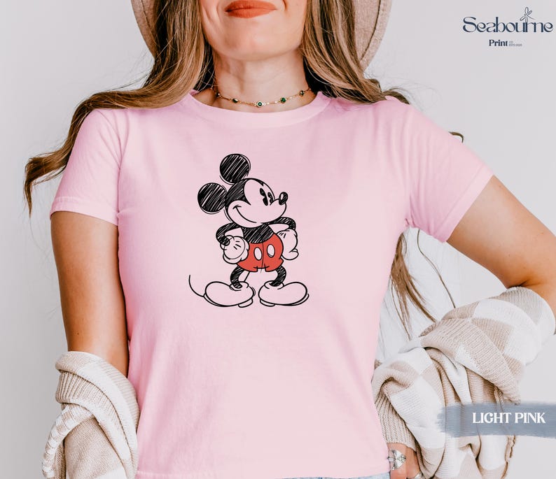 May include: Light pink t-shirt featuring a black and white sketch of Mickey Mouse with red shorts. The shirt has short sleeves and a crew neck. The text "LIGHT PINK" is visible in the bottom right corner.