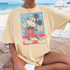 K&ouml;nnte beinhalten: Ein hellgelbes Kurzarm-T-Shirt mit einer Grafik von Mickey Mouse, die einen Daumen hoch zeigt. Das Design enth&auml;lt den Text USA, wei&szlig;e Sterne auf blauem Grund und rote horizontale Streifen. Das T-Shirt hat einen Rundhalsausschnitt.