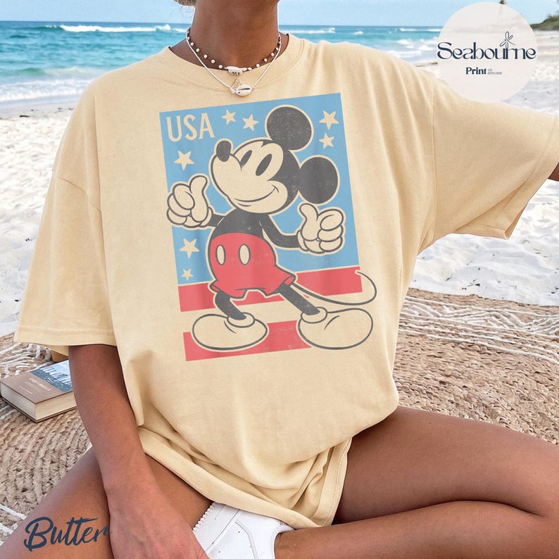 K&ouml;nnte beinhalten: Ein hellgelbes Kurzarm-T-Shirt mit einer Grafik von Mickey Mouse, die einen Daumen hoch zeigt. Das Design enth&auml;lt den Text USA, wei&szlig;e Sterne auf blauem Grund und rote horizontale Streifen. Das T-Shirt hat einen Rundhalsausschnitt.