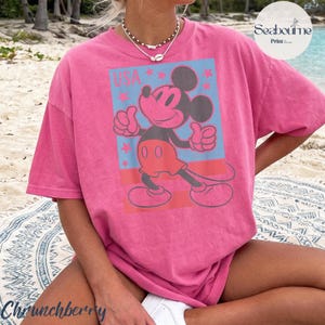 K&ouml;nnte beinhalten: Ein rosafarbenes Kurzarm-T-Shirt mit einer Grafik von Mickey Mouse in roter Kleidung vor blauem Hintergrund, roten Sternen und dem Text USA. Das Motiv ist mittig auf der Brust platziert. Das Shirt hat einen Rundhalsausschnitt und eine lockere Passform.