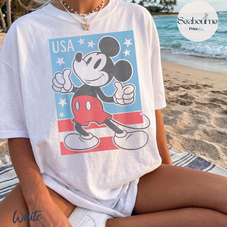 K&ouml;nnte beinhalten: Ein wei&szlig;es Kurzarm-T-Shirt mit einer Grafik von Mickey Mouse, die den Daumen hoch zeigt. Das Design enth&auml;lt den Text USA, wei&szlig;e Sterne auf hellblauem Grund und rote horizontale Streifen. Die Grafik hat ein abgenutztes Vintage-Finish.